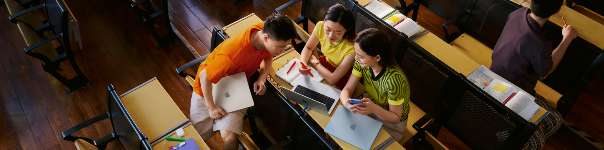 大学教室的俯拍照片，展现三个学生人手一台 MacBook，其中一位女生的 MacBook 打开放在课桌上，他们正围着屏幕进行讨论。